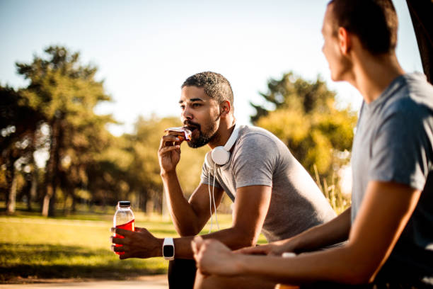 Active young male taking a bite out of an protein bar and hydrating himself with a sport drink next to his friend stock photo