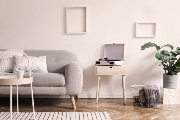 istock Stylish scandinavian white interior with design sofa, tropical plant, pillows, blanket, gramophone and mock up photo frames. White background walls, brown wooden parquet and modern triangle lamp. 1146020157