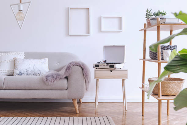 istock Stylish and cozy scandinavian white interior with design sofa, bookstand with accessories, pillows, blanket, gramophone and mock up photo frames. White background walls and modern triangle lamp. 1146020179