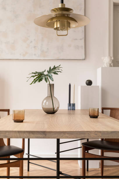 istock Eclectic and elegant dining room interior with design sharing table, chairs, gold pedant lamp, abstract paintings and stylish accessories. Tropical leafs in vase. Eclectic decor. Brown wooden parquet. 1146020407