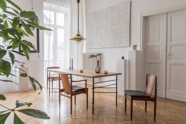 istock Stylish and modern dining room interior with design sharing table, chairs, gold pendant lamp, abstract paintings and elegant accessories. Tropical leafs in vase. Eclectic decor. Brown wooden parquet. 1146020408