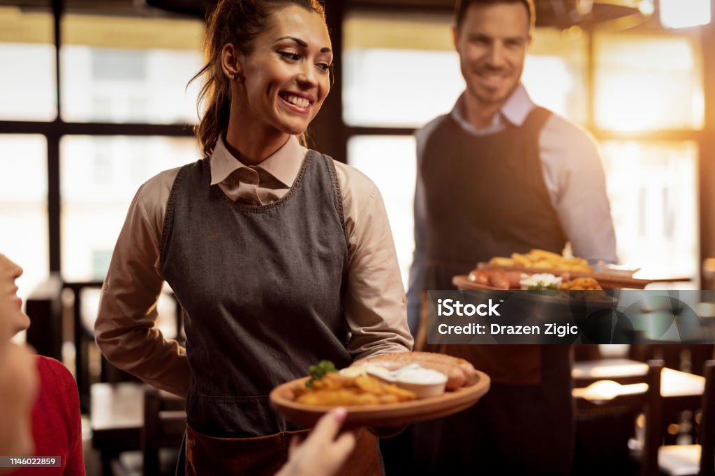 Happy waiters serving food to their guests in a restaurant. Two waiters serving lunch and brining food to their gusts in a tavern. Focus is on happy waitress. Meat Stock Photo