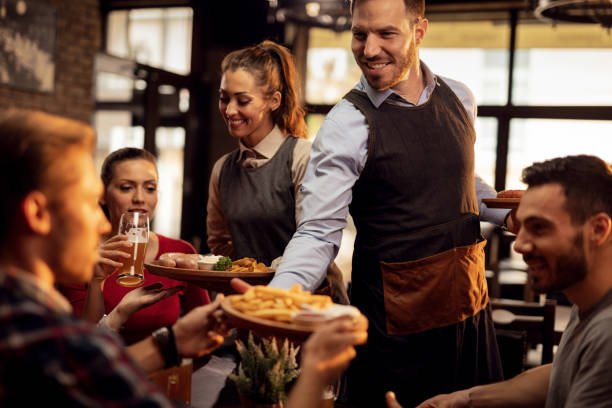 Young waiters serving lunch to group of friends in a pub. Happy waiters bringing food at the table and serving group of friends in a restaurant. waiter stock pictures, royalty-free photos & images