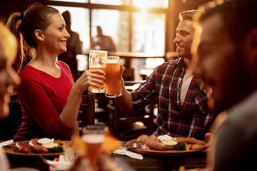Young smiling couple celebrating and toasting with beer while having lunch with friends in a pub.