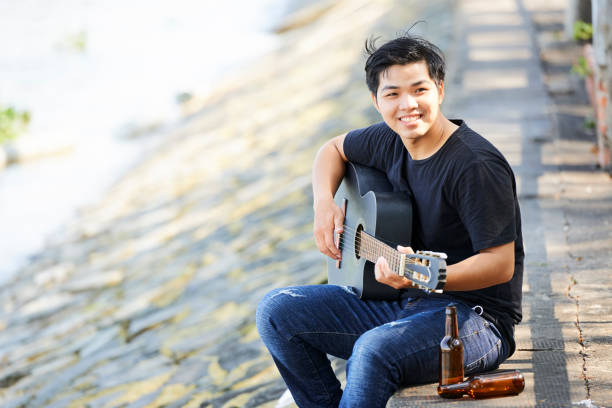 Asian young man resting outdoors with empty bottles of beer playing guitar and smiling