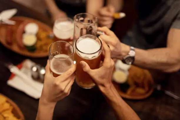 Group of unrecognizable friends  celebrating and toasting with beer while having lunch in a tavern.