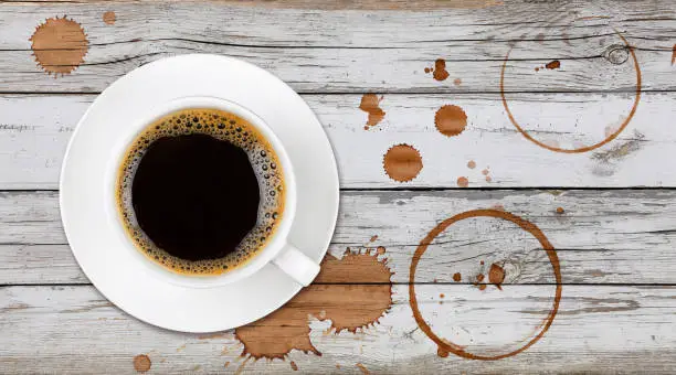 Brown cup full of black coffee on saucer, with ring coffee stains and drops on white painted wooden table background, elevated top view, directly above