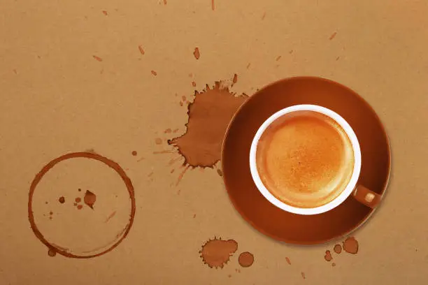 Brown cup full of espresso coffee on saucer, with ring coffee stains and drops on brown paper parchment background, elevated top view, directly above