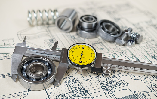Analog metallic measuring instrument with yellow round dial. Group of steel components on document. Precise calliper. Selective focus