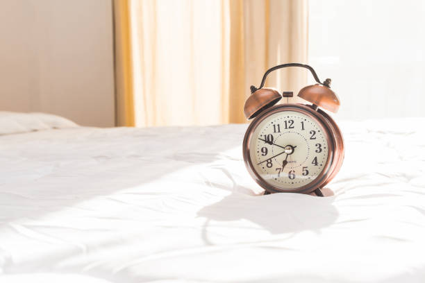 Alarm clock in white bed with soft sunlight at morning stock photo