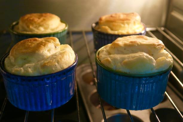Image of four homemade cheese souffles made with whisked egg whites and herbs, rising in hot oven for dinner party, delicious golden brown cheese souffle starter appetiser cooking in round blue China ramekin dishes, home baking photo on oven grill shelf Stock photo showing four homemade cheese souffles, which are pictured rising nicely and going a golden brown colour on the top, in hot oven for dinner party starter dish / appetiser, ready to be served with a leafy green salad. Made with whisked egg whites (beaten like a meringue mixture) and herbs, these delicious cheese souffle dishes are cooking in attractive round, blue and green glazed China ramekin dishes, with this home baking photo also featuring an oven grill shelf and blurred non-stick muffin cupcake tray in the background. appetiser stock pictures, royalty-free photos & images