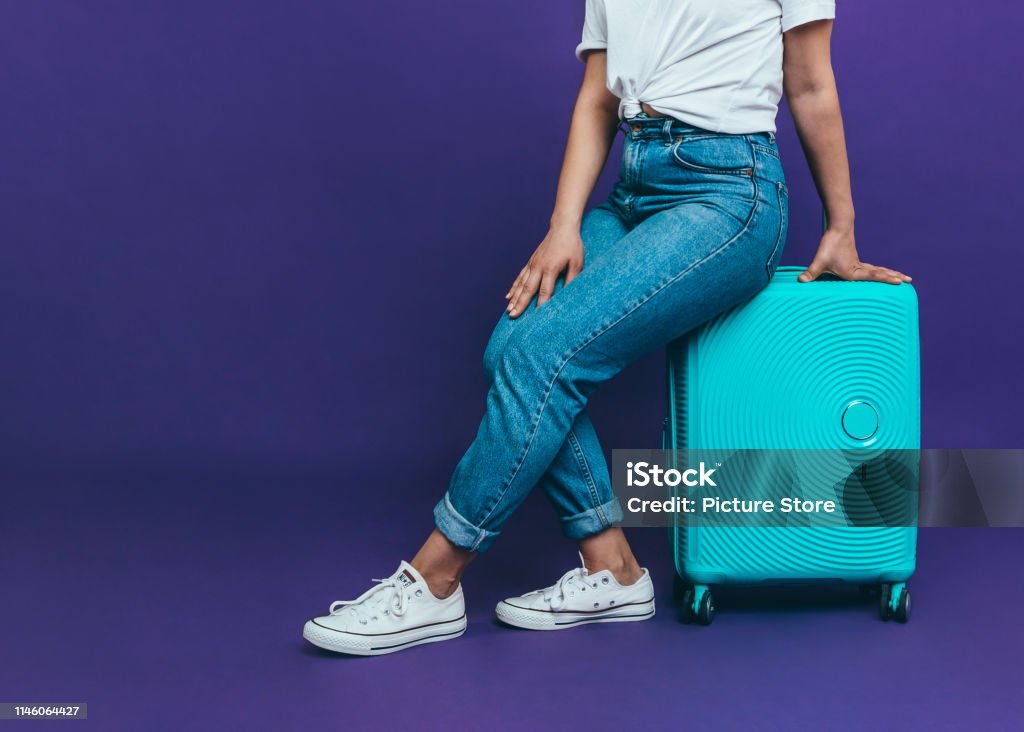 Travel bag Close-up photo A young woman in blue jeans is sitting on a blue suitcase on a purple background Adult Stock Photo