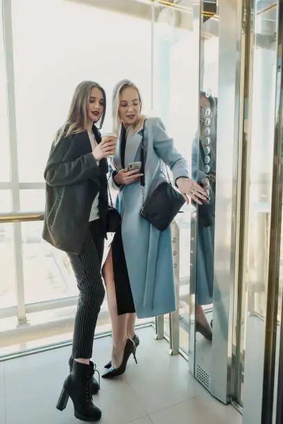 Photo of Two beautiful girls, a blonde and a brunette, are wearing a coat in a glass elevator with a window. Shopping center or office in the city.