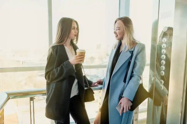 Photo of Two beautiful girls, a blonde and a brunette, are wearing a coat in a glass elevator with a window. Shopping center or office in the city.