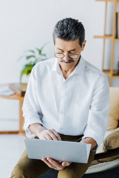 istock pensioner with mustache and glasses using laptop while sitting on sofa 1146072802
