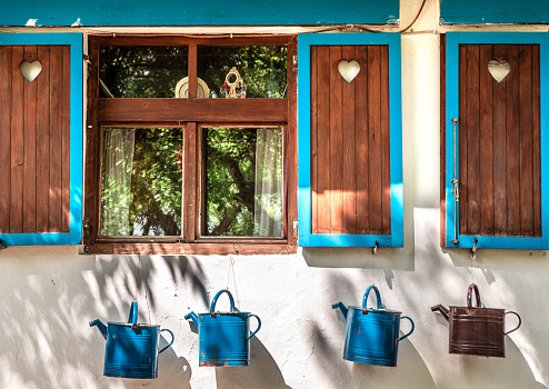 Cute old village etno house windows with wooden blinds with heart shaped holes. Blue watering cans as decoration underneath.