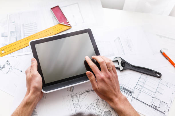 istock Man hand working with tablet and blueprint on architectural project at home while relaxing on couch. Back view of latin architect reading plan layout on digital tablet. 1146106911