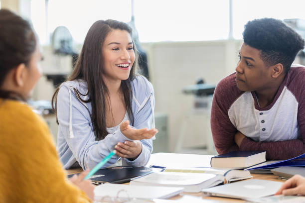 Teen girls debates with peers in class A cheerful teen girl gestures as she sits at a table in her classroom and debates with peers. debate stock pictures, royalty-free photos & images