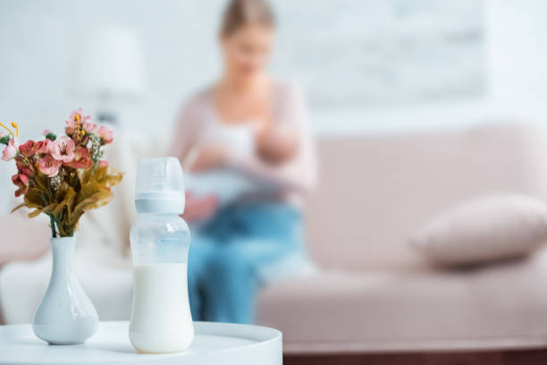 крупным планом вид детской бутылки с молоком, цветы в вазе и мать грудного вскармливания ребенка позади дома - milk bottle стоко�вые фото и изображения