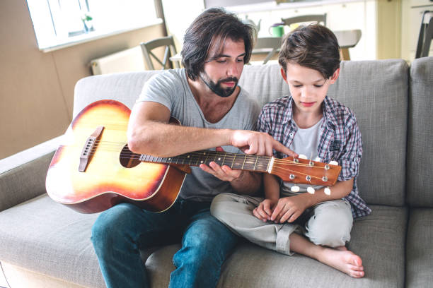 istock Man is istting on sofa with his son. He holds guitar. Man is pointing on strings. Boy looks at it . They are concentrated. 1146124022
