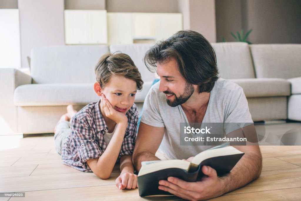 Boys are lying on the floor. Man holds book and look at son. He smiles to him. Boy is shy. He hols hand on face. Boys are lying on the floor. Man holds book and look at son. He smiles to him. Boy is shy. He hols hand on face Adult Stock Photo