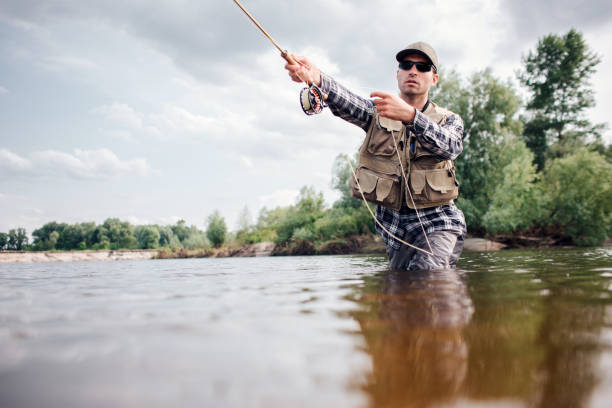 fischer in aktion. guy wirft löffel fliegenstange in wasser und hält einen teil davon in der hand. er schaut geradeaus. der mensch trägt spezielle schutzkleidung. - fischereiindustrie stock-fotos und bilder