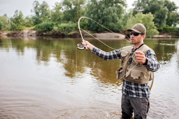 un pescatore serio si trova su un superficiale agitando con la pesca a mosca. inoltre ha un cucchiaio in altra mano. il tizio guarda a sinistra. è concentrato. - lure loc foto e immagini stock