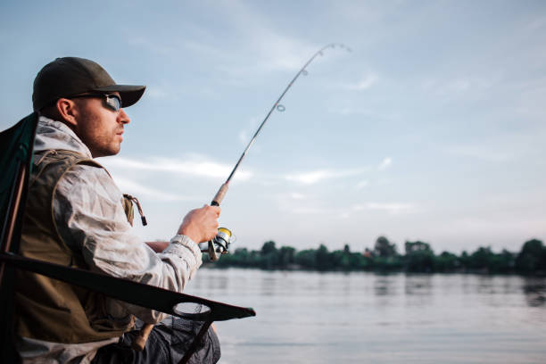thoughtful guy is sitting at the edge of water and looking to the right. he holds fly rod in hands. it is evening and chilly outside. - animal catch of fish catching sport imagens e fotografias de stock