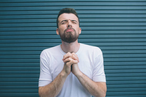 istock Young man is standing and holding hands in pray position. He is keeping eyes closed. Guy is holding his head up. Isolated on striped and blue background. 1146124970
