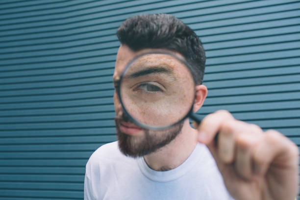 istock Suspicious guy is standing and looking through loup on camera. He is showing one eye and holding this thing in one hand. Isolated on striped and blue background. 1146125017