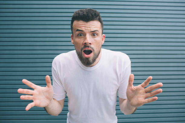 istock Guy in white shirt is standing and looking at camera. He is amazed very much. Guy has opened his mouth and holding hands in shot. Man's brows are raised up. Isolated on striped and blue background. 1146125025