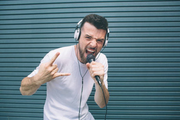 bello tizio è in piedi e in posa sulla macchina fotografica. sta urlando al microfono e mostrando il segno bello. l'uomo indossa le cuffie. sta ascoltando musica. isolato su sfondo a strisce e blu. - instrumental hip hop immagine foto e immagini stock