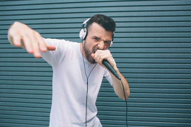 istock Cool and awesome man is reading rap in microphone. He is showing his hand and looking at camera. Guy is singing in mic and wears headphones. Isolated on striped and blue background. 1146125042