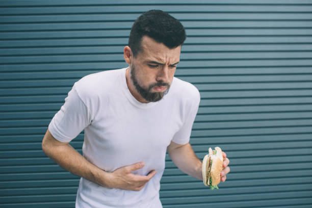 Guy is sick. He is holding burger in hand and looking down. Man is going to vomit. Isolated on striped and blue background. Guy is sick. He is holding burger in hand and looking down. Man is going to vomit. Isolated on striped and blue background puke green color stock pictures, royalty-free photos & images