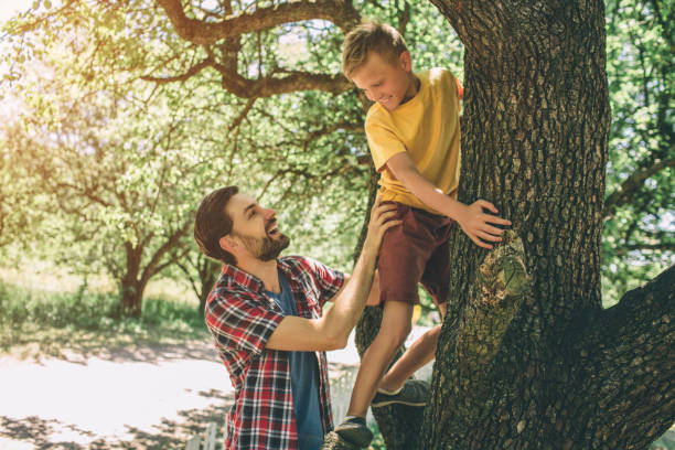 istock Bearded guy is standing near tree and supporting his son. Boy is climbing up. Child has put his hands on tree and looking at his father. They are smiling. 1146125265