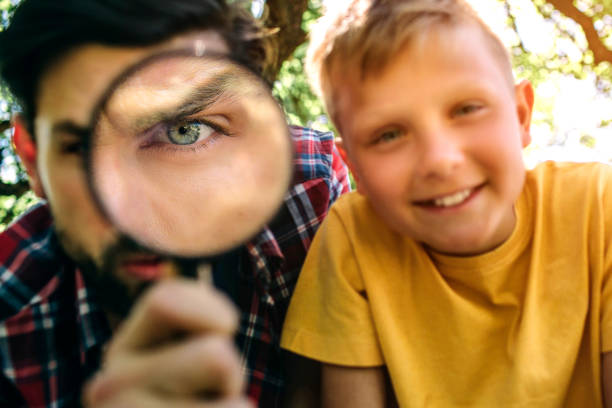 istock Bearded guy is looking on camera through loop. Oni one eye is zoomed in. Adult looks serious and suspicious. His son is looking on camera and smiling. He is happy. 1146125281