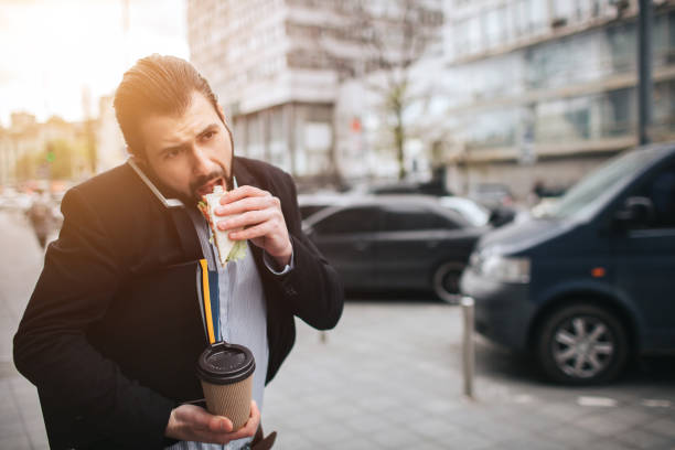 Busy man is in a hurry, he does not have time, he is going to eat snack on the go. Worker eating, drinking coffee, talking on the phone, at the same time. Businessman doing multiple tasks. Multitasking business person. stock photo