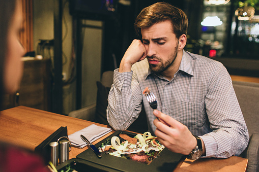 Pictue of guy sitting together with his girlfriend and eating some food they have ordered. Man has found some meat on the plate. He is looking to the piece of meat with suspicious sight