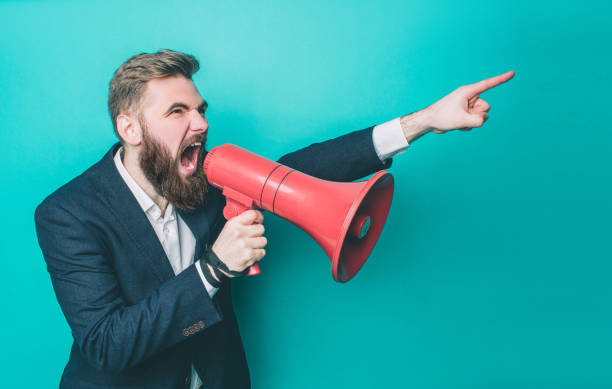 istock Serious guy is looking straight forward. He is pointing and commanding. Man is screaming in the loudspeaker. Isolated on blue background. 1146126426