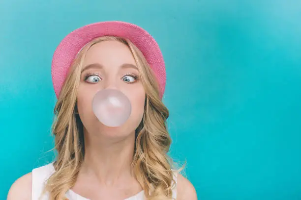 Photo of Funny girl is inflating the ball from chewing gum. Also she is looking on this ball in a funny position. Isolated on blue background.