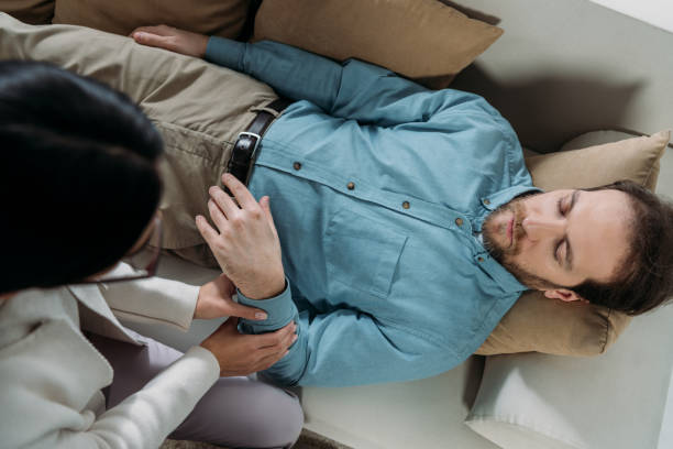 overhead view of hypnosis holding wrist of bearded man with closed eyes lying on couch overhead view of hypnosis holding wrist of bearded man with closed eyes lying on couch hypnosis stock pictures, royalty-free photos & images