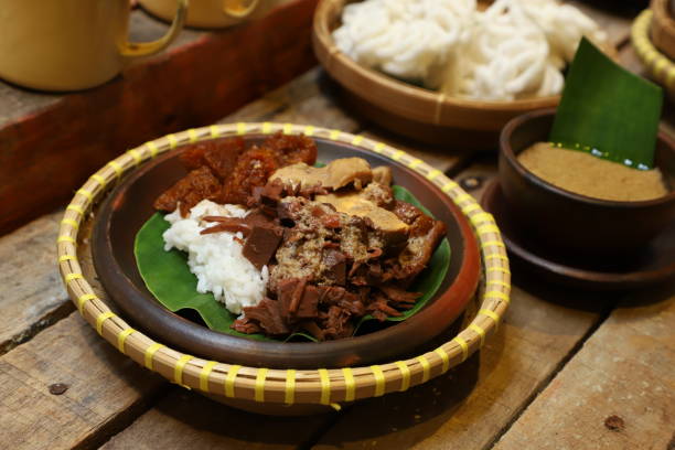 Nasi Gudeg, the Traditional Javanese Rice Dish with Jackfruit Stew, White Chicken Curry and Spicy Cattle Skin Stew Nasi Gudeg, a traditional Javanese meal, consists of steamed rice topped with jackfruit stew (Gudeg), white chicken curry, and spicy stew of cattle skin crackers. The jackfruit stew is then drizzled with thick sauce from the chicken curry. Plated on an earthenware plate lined with banana leaf. Arranged on a rustic wooden table together with some crackers. gudeg stock pictures, royalty-free photos & images