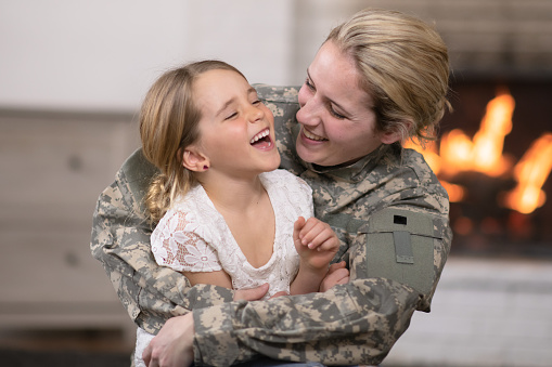 A mother and her daughter are hugging in their living room. The mother is wearing a military uniform while the girl has a giant smile on her face. She is so excited that her mom is home.