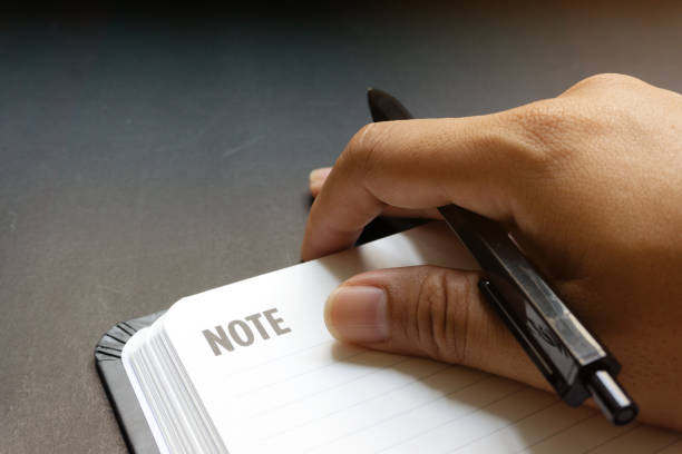 Close up human hands with black pen writing something on blank line notebook with black desk background in dramatic lighting tone.Concept for business, planning or diary.Great for any use. cierre las manos humanas con un bolígrafo negro escribiendo algo sobre un cuaderno de línea en blanco con fondo de escritorio negro en un tono de iluminación dramático. concepto de negocio, planificación o diario. ideal para cualquier uso.