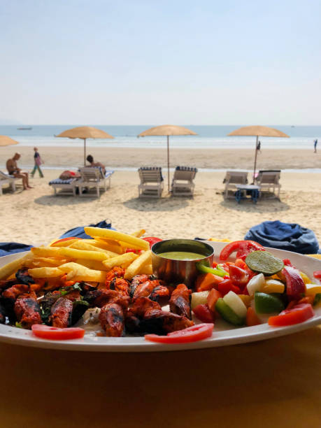 Image of alfresco dining prawns, chips and tomatoes at Palolem Beach, Goa, India seafood restaurant with barbecued king prawn kebabs, chips / French fries, green chutney and salad garnish with red peppers, seaside sun lounger, beach parasols in background Stock photo showing a scenic alfresco dining spot with a large white oval plate, complete with prawns and chips at Palolem Beach, Goa, India. This seaside restaurant is famous for its barbecued king prawn kebabs, which come served with crunchy chunky chips / French fries, a silver dish of green chutney (coriander and mint) and a colourful salad garnish with red peppers / chillies, slices of tomato, sun loungers, parasols, sunbathers and sea in background, as well as the golden sand of the gorgeous tropical paradise that is Palolem Beach itself. palolem beach stock pictures, royalty-free photos & images