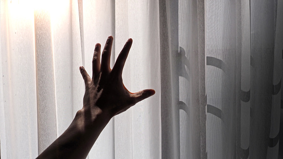 Silhouette of a male hand reaching out to the white curtain with morning sunlight effect.