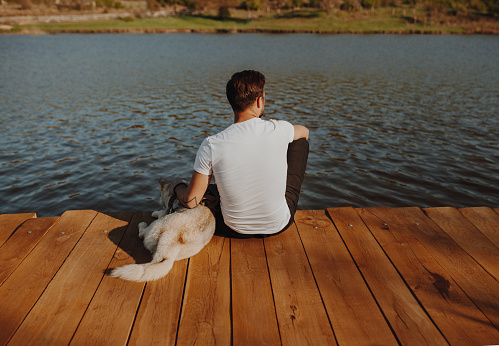 Back view of man sitting on wooden pier above lake with dog near and looking away