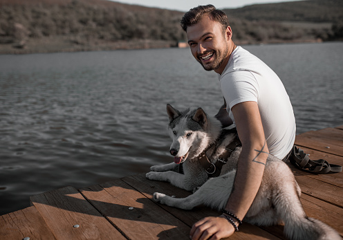 Side view of cheerful hipster with beautiful Husky dog sitting on wooden pier smiling at camera