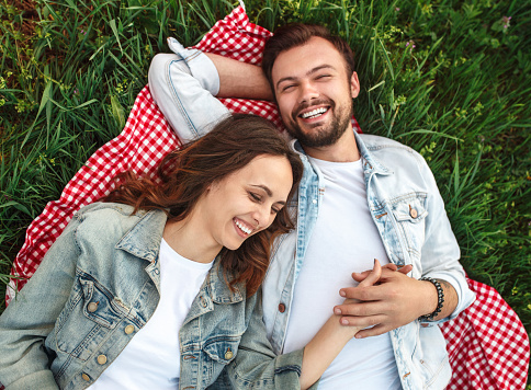 From above shot of young man and woman smiling while lying together on checkered blanket on green grass in nature
