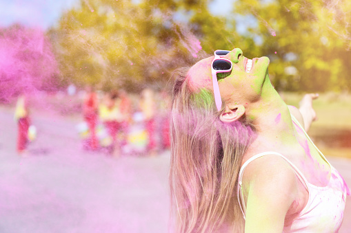 Woman loving the color festival, laughing and splashing hair in pink colored powder. Young candid girl with face paint wearing pink sunglasses and enjoying the event.